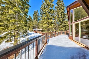 A snow-covered deck with a wooden railing, surrounded by tall pine trees.