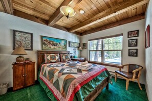 A bedroom with a wooden ceiling, a large bed with a quilt, a wooden chair, and a window with a view of trees.