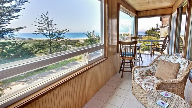 Een balkon met een rieten bank en stoelen, grote ramen met uitzicht op zee en groen.
