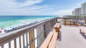 Blick auf einen Strand von einem Balkon mit Holzgeländern, einer Holzdielen-Terrasse und Meer, sowie Personen am Strand.