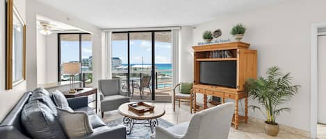 A spacious living area with a sofa, chairs, a coffee table, a TV stand, and a view of the ocean.