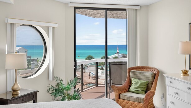 Ein Hotelzimmer mit großem Fenster und Blick auf Strand und Meer, ein Korbstuhl mit Kissen, ein Nachttisch mit Lampe sowie eine weiße Kommode.