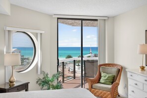 Quarto de hotel com uma grande janela que oferece vista para a praia e o oceano, uma cadeira de vime com almofada, uma mesa de cabeceira com luminária e um aparador branco.
