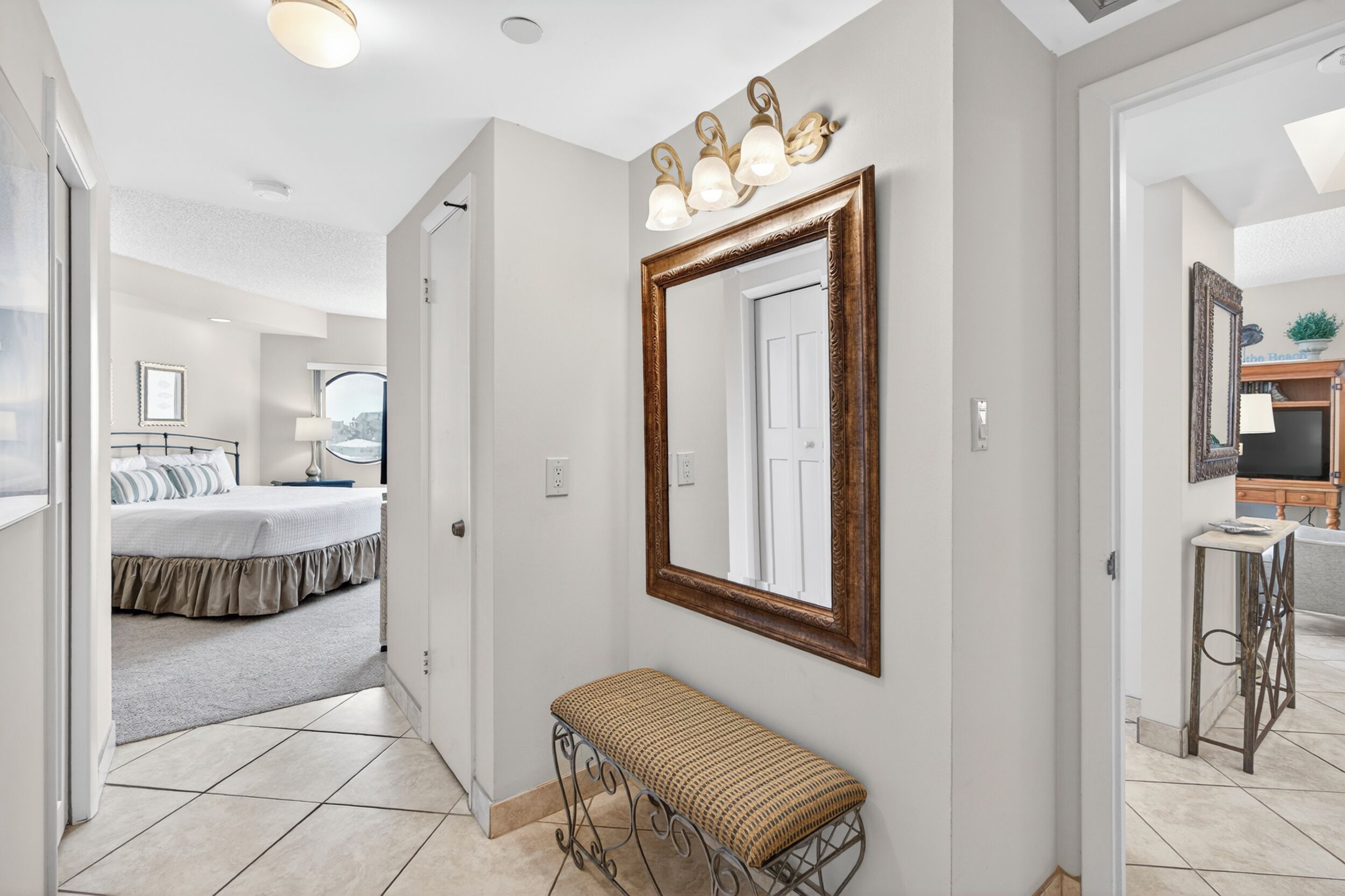 A hallway with a large mirror, a bench, and a door leading to a bedroom with a bed and a window.