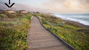 Una pasarela de madera que conduce a una playa con vistas al océano, rodeada de vegetación y algunos edificios a lo lejos.