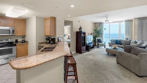 A modern kitchen with granite countertops, a built-in microwave, and a stove. There is a dining area with a bar stool and a large window overlooking the ocean. The living room features a gray sofa and a television. - 0602 - Modern Gulf Front Oasis (Panama City Beach)