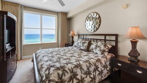 A bedroom with a bed, two bedside tables, a ceiling fan, a large window with a view of the ocean, and a decorative mirror on the wall. - 0602 - Modern Gulf Front Oasis (Panama City Beach)