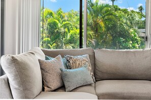 A beige sofa with patterned cushions in a room with large windows overlooking a tropical garden.