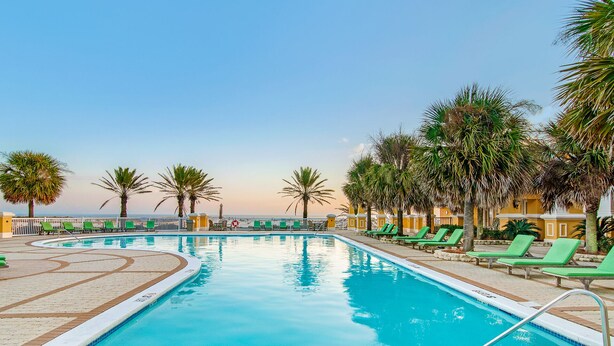 Zona de piscina con vista despejada al océano, rodeada de palmeras y sillones de descanso verdes.