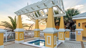 Zona de piscina con una pérgola, palmeras y una puesta de sol al fondo.