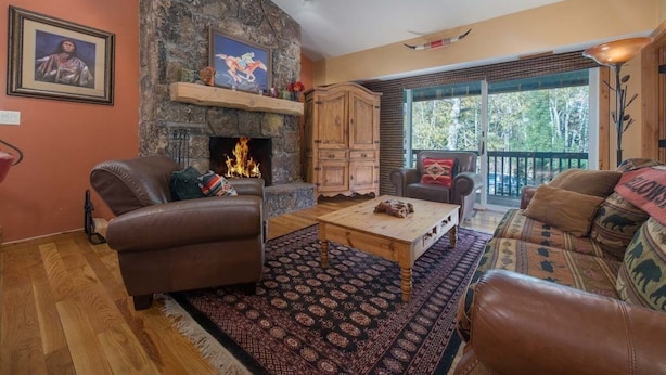 Una sala de estar con chimenea de piedra, sofás de cuero, una mesa de centro de madera y un ventanal con vistas a árboles.