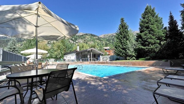 Un espace aménagé au bord de la piscine, avec une table et des chaises, un grand parasol et une vue sur les montagnes et les arbres.