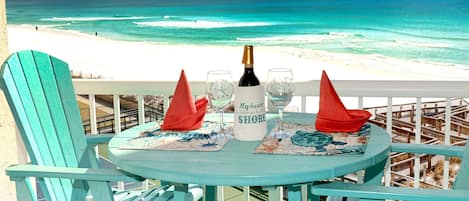 A turquoise table set with a bottle, glasses, and a placemat with a coastal design, overlooking a beach.