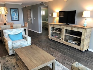 Uma sala de estar com uma mesa de centro de madeira, um sofá de vime com almofada azul, um rack de madeira com uma TV de tela plana e uma luminária fixada na parede.
