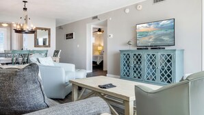 A living room with a TV, a dining area with chairs, a sofa, and a wooden table. - 🌅 Islander #403 – Sunrise Views & Front-Row Beach Action! 🌊🏄‍♂️ (Destin)