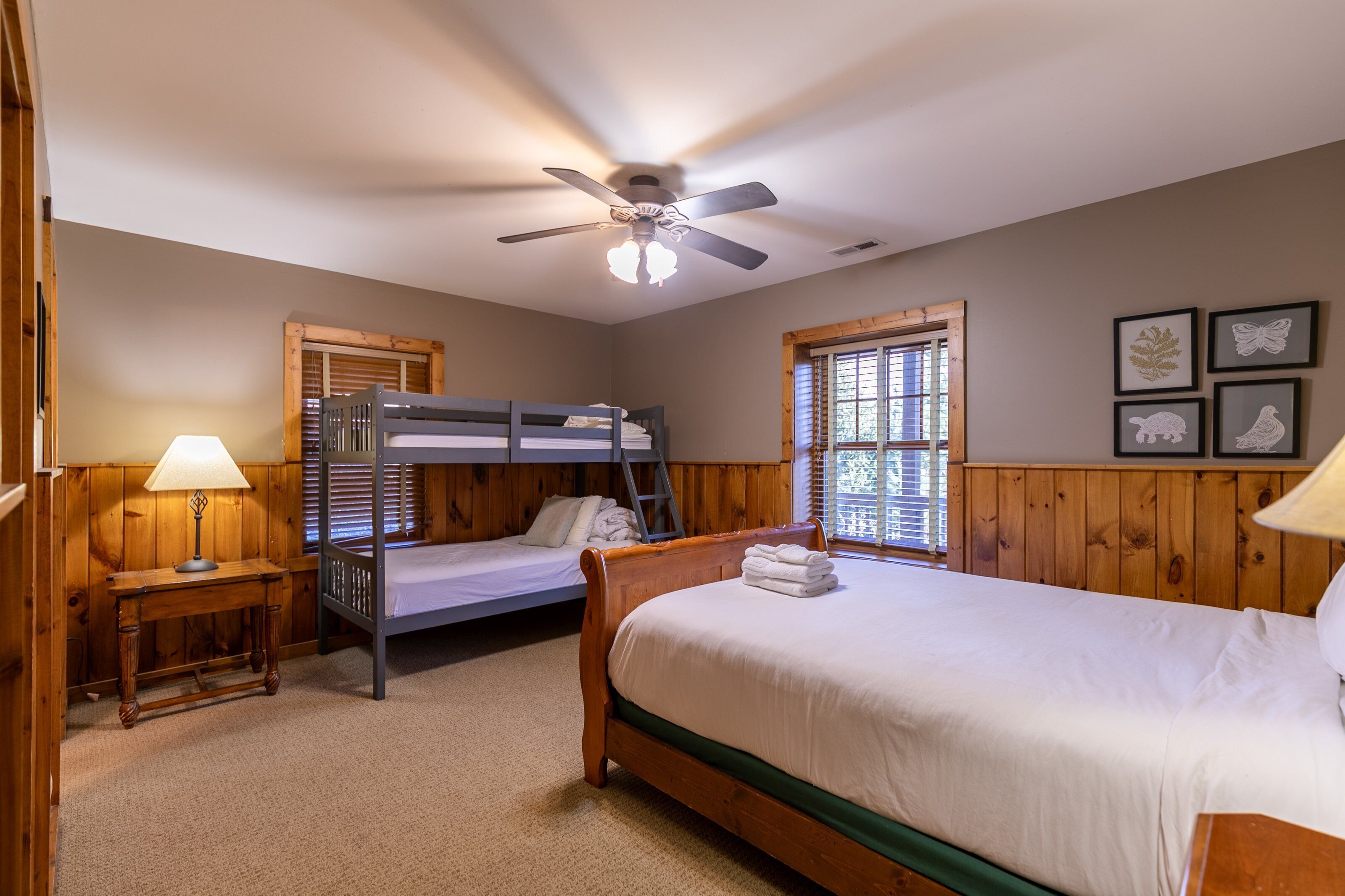 Une chambre avec un lit superposé, un lit simple, un ventilateur de plafond, une fenêtre avec des stores et des tableaux encadrés accrochés au mur.