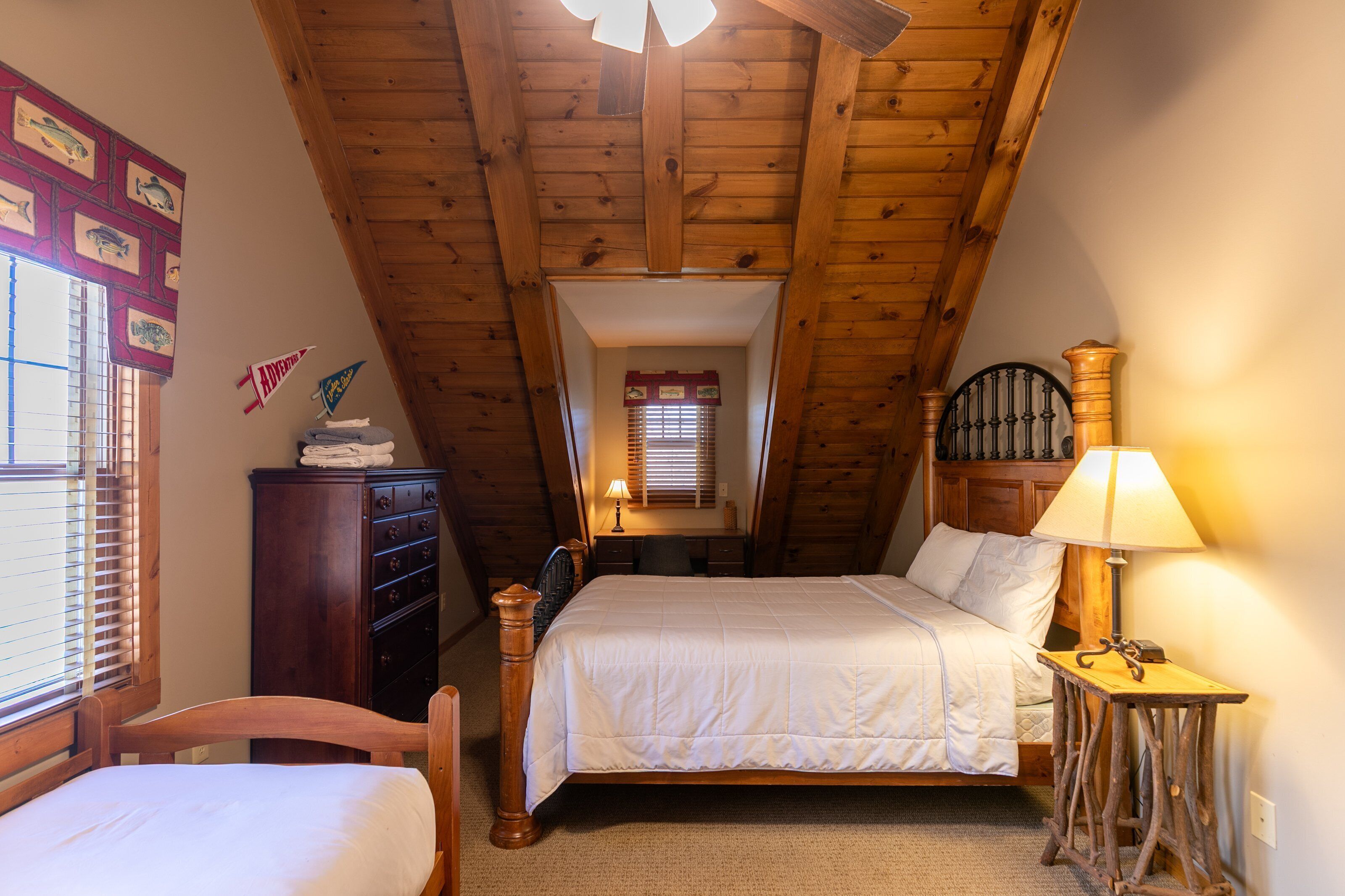 A bedroom with a sloped ceiling, wooden headboard, a bed with white bedding, a desk with a lamp, and a dresser.
