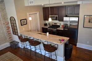 A kitchen with a central island, bar stools, stainless steel appliances, and a brick wall. - 1890-1930 Historic Politics Themed condo, 2 blocks from French Quarter (New Orleans)