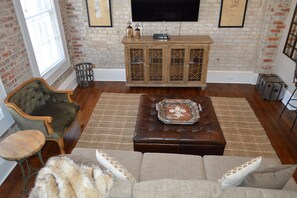 A living room with a brick wall, a television mounted above a wooden cabinet, a leather sofa, and a patterned rug. - 1890-1930 Historic Politics Themed condo, 2 blocks from French Quarter (New Orleans)