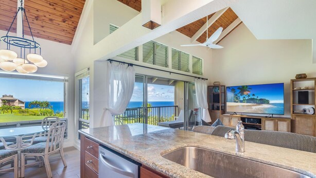 A modern kitchen with a large island, a dining area with chairs, a built-in dishwasher, and a view of the ocean. - Pali Ke Kua 245 .. Updated Oceanfront Condo, Views are Expansive! (Princeville)