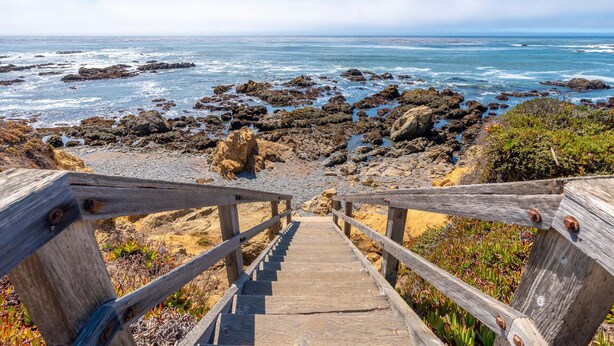 Un escalier en bois mène à une plage rocheuse d’où l’on peut admirer l’océan.