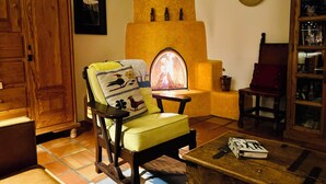 A cozy living room with a fireplace, a wooden cabinet, a chair with a patterned cushion, and a coffee table with magazines.