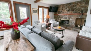 A living room with a stone fireplace, grey sofas, a wooden coffee table, and a vase of red flowers. - JHRL - Rendezvous B4, Comfortable Slope-side Condo, Hot tub next door (Teton Village)