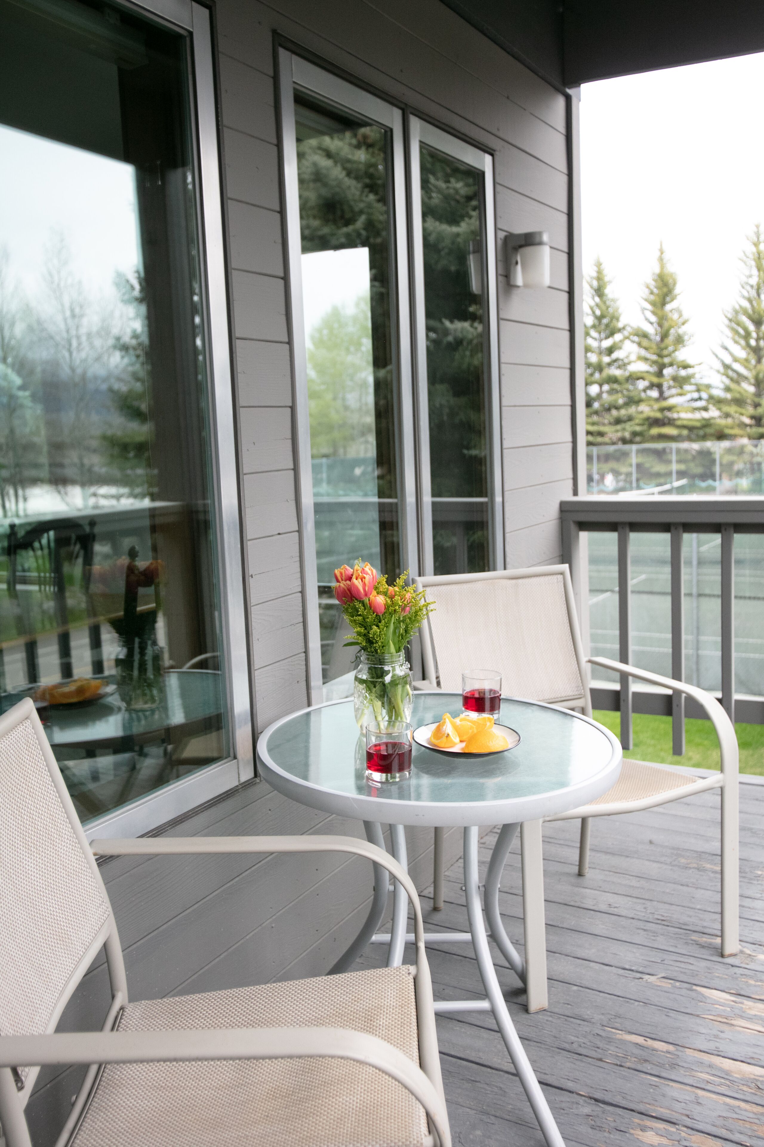 A glass table with chairs on a wooden deck, a vase with flowers, and a glass of red wine.
