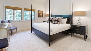 A four-poster bed with a dark headboard, a bedside table with drawers, and a lamp. There is a window with a view of trees, a chair, and a desk with a lamp. - JHRL - Wind River #2, Well-Appointed and Well-Located - 1 minute from Ski Access (Teton Village)