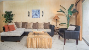 Een woonkamer met een hoekbank, een salontafel, twee fauteuils, een vloerlamp en een grote plant.