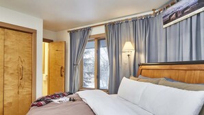 A hotel room with a wooden door, a bed with white and patterned bedding, a window with curtains, and a wall-mounted lamp. - JHRL - Rendezvous D4, Great location for families (Teton Village)
