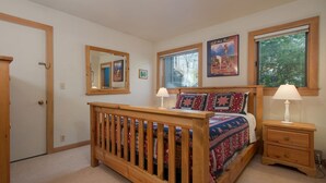 A bedroom with a wooden bed, two bedside tables, a window with blinds, a mirror, and a door.