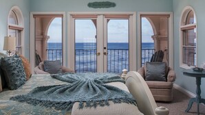 A bedroom with a large bed, two chairs, a small table, and a view of the ocean through a set of doors.