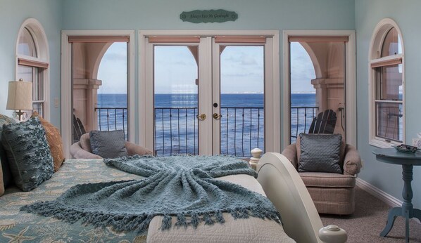 Een slaapkamer met een groot bed, twee stoelen, een klein tafeltje en uitzicht op de oceaan door een reeks deuren.