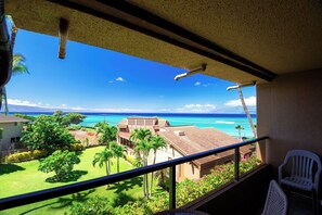 A balcony with a view of a tropical landscape, including a lawn, palm trees, and a house. - OCEAN VIEW 1 Bedroom condo - Steps to the beach! Kahana Villa F406 (Lahaina)
