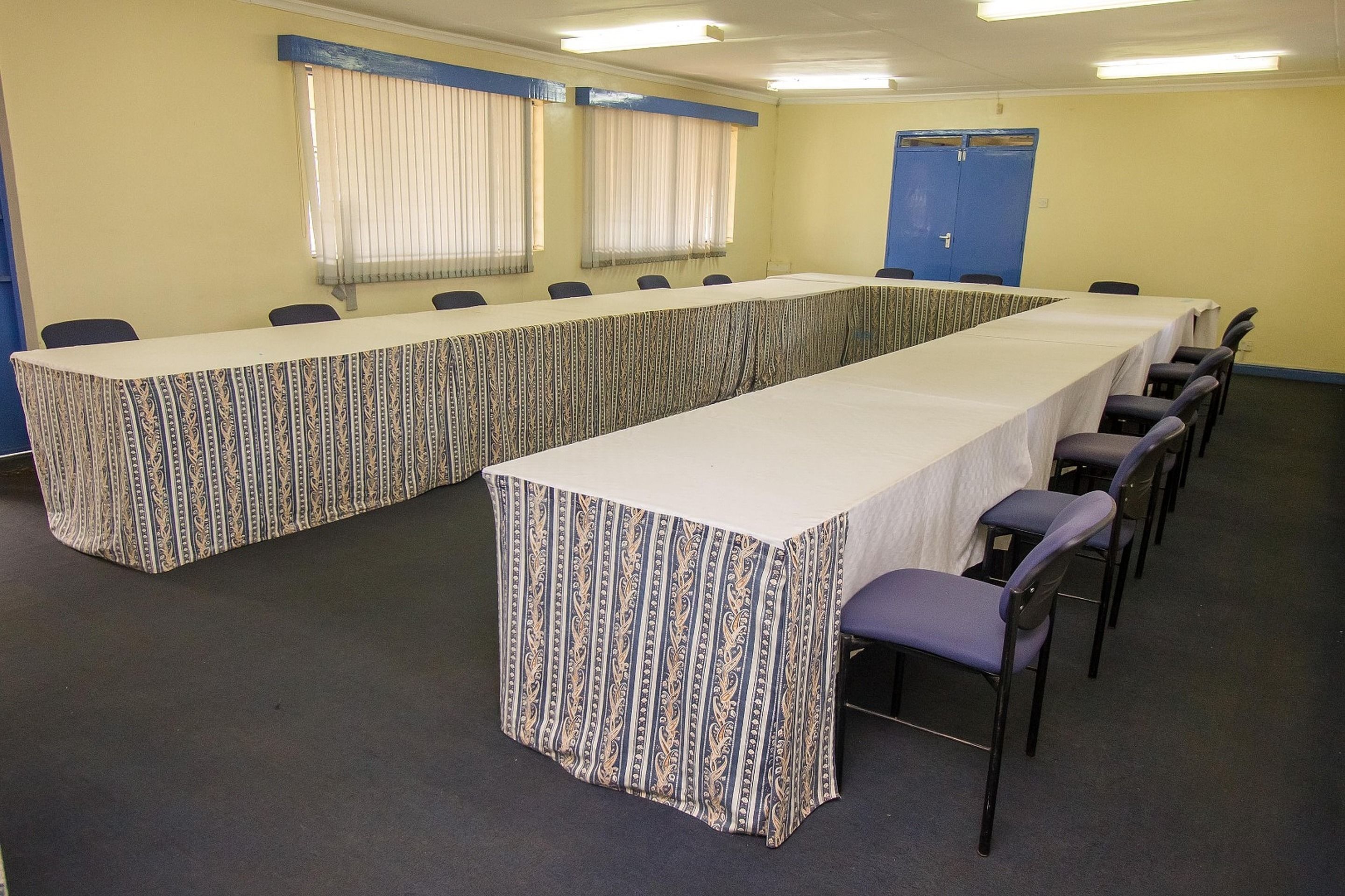 Une salle de conférence avec une longue table et des chaises, une porte bleue et des fenêtres avec des rideaux.