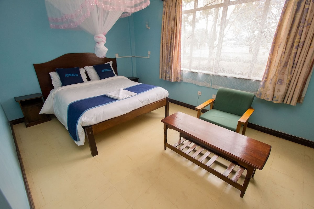 Un dormitorio con una cama, un banco de madera, una silla verde y una ventana con cortinas.