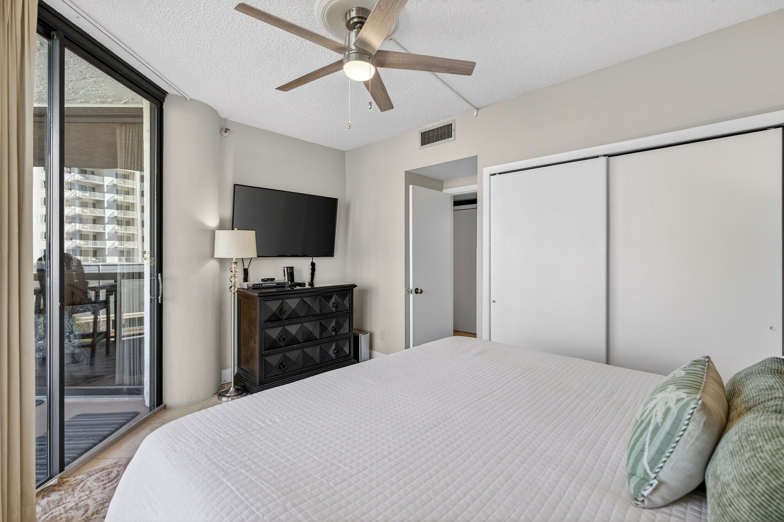 Une chambre avec un grand lit, un ventilateur de plafond, un téléviseur fixé au mur et une porte coulissante donnant sur un balcon.