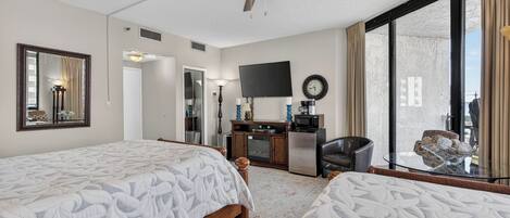 Habitación de hotel con dos camas, ventilador de techo, televisor, escritorio con microondas y un ventanal grande con cortinas.