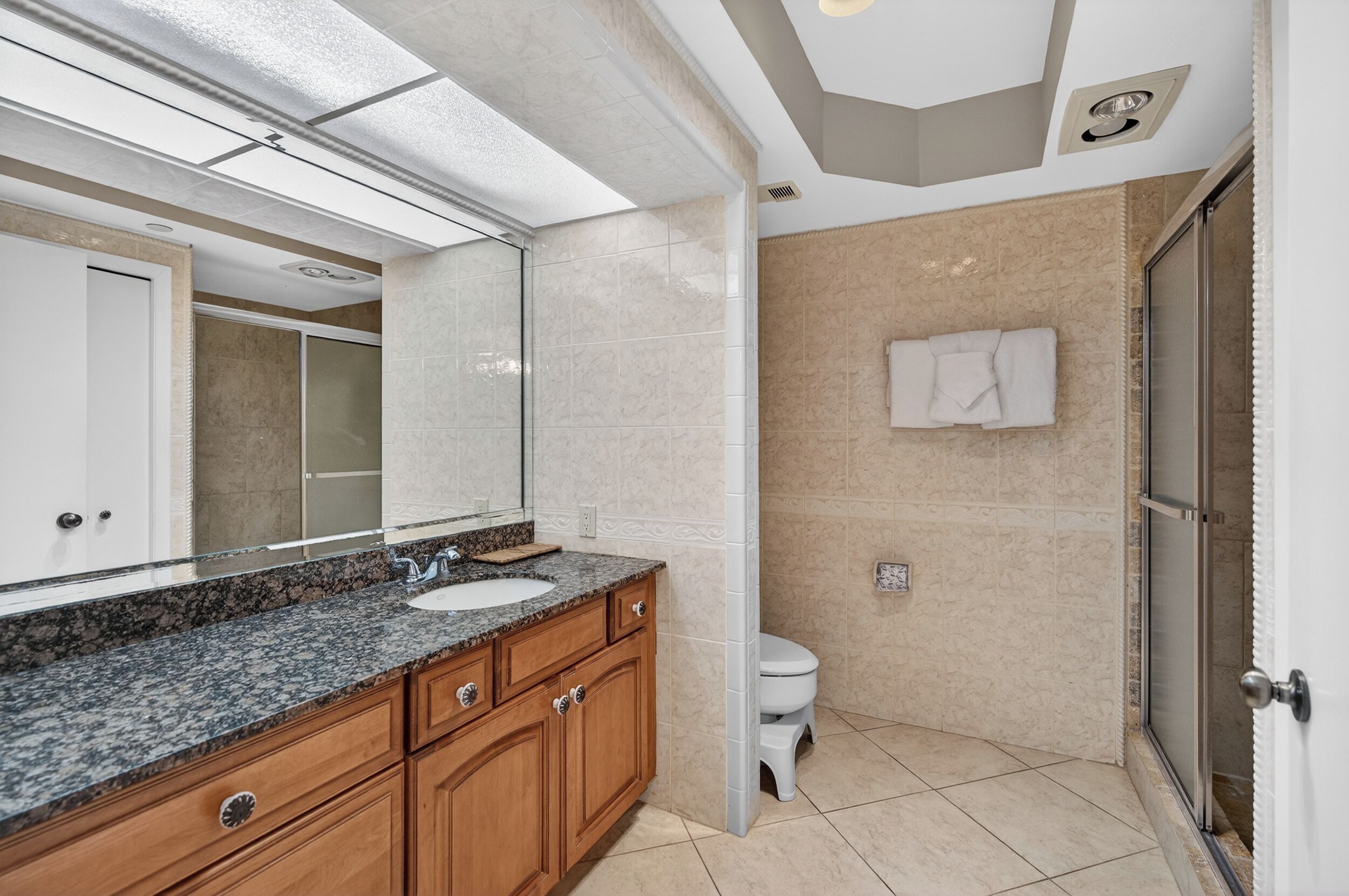 A modern bathroom with a granite countertop sink, wooden cabinets, a large mirror, a toilet, and a shower area.