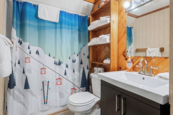 A bathroom with a mountain-themed shower curtain, a white toilet, a wooden shelf with towels, a sink with a mirror, and a bottle of hand soap.