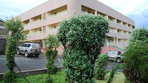 A multi-story building with a flat roof and balconies. There are vehicles parked in the driveway.