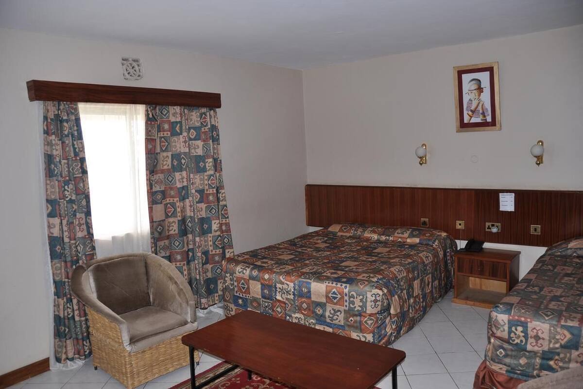 A room with two beds, a chair, a wooden headboard, patterned curtains, and a wall-mounted picture.