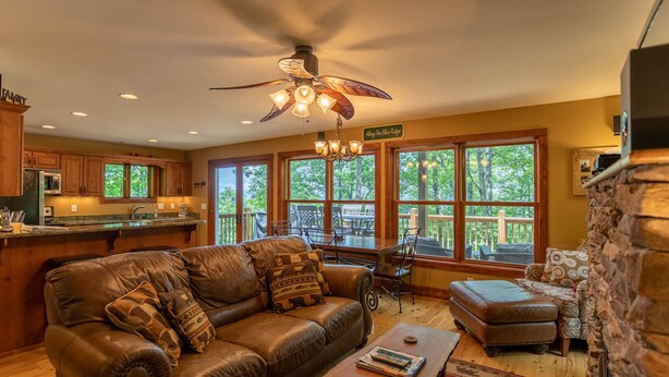 A spacious living room with a leather sofa, a wooden coffee table, and a stone fireplace. There is a kitchen area with wooden cabinets and a large window overlooking a deck. - A Peak of Heaven 1 mi from Blue Ridge Parkway very private ping pong table (Boone)