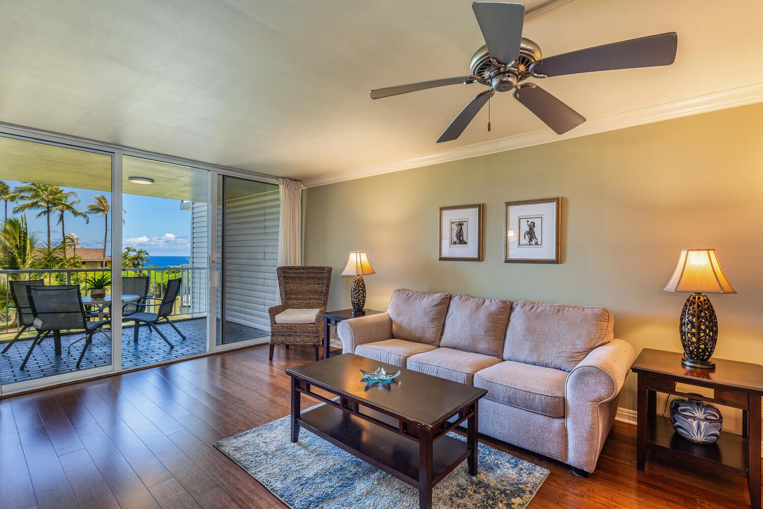 Ein geräumiges Wohnzimmer mit einer großen Schiebeglaswand, die auf einen Balkon mit Meerblick führt. Ein Deckenventilator, ein Sofa, zwei Beistelltische mit Lampen und ein Couchtisch sind ebenfalls vorhanden.