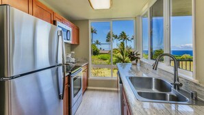 A kitchen with stainless steel appliances, wooden cabinets, a granite countertop, and a large window offering a view of palm trees and the ocean. - CLIFFS 4-207,  SPECTACULAR VIEWS - 2 PRIVATE LANAIS AT THIS REMODELED CONDO (Princeville)