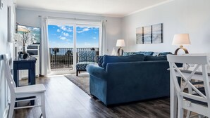 A modern living room with a blue sofa, a white chair, and a dining table with two lamps. A large window offers a view of the sky and a balcony. - 🌅 Islander #304 – Affordable Beach Getaway with Amazing Sunrises! 🌊✨ (Destin)