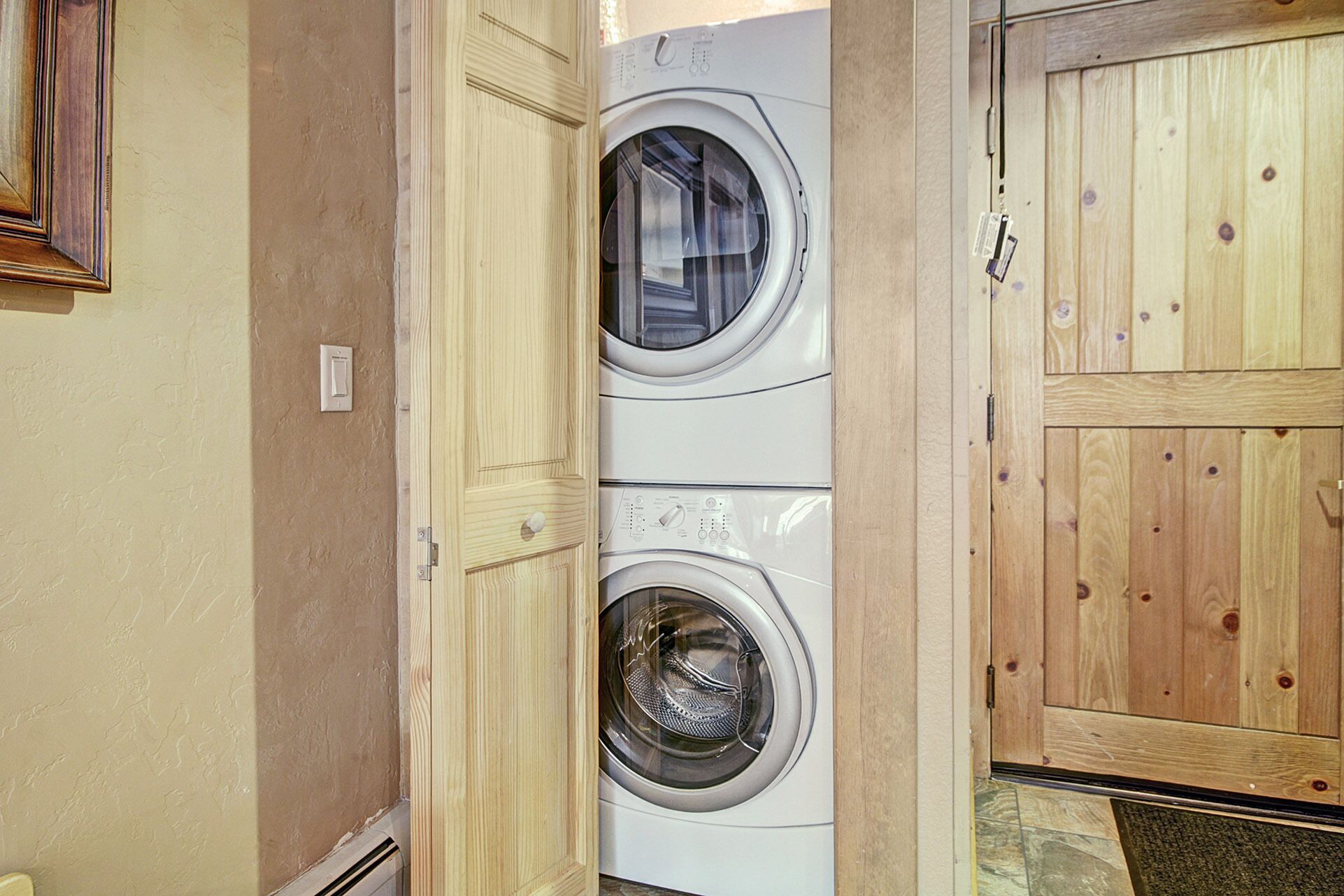 A laundry area with two stacked front-loading washing machines, a wooden door, and a light switch on the wall.