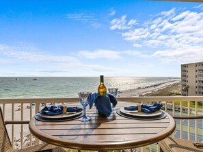 A table set for two with a bottle of wine, two wine glasses, and a blue cloth on a balcony overlooking a beach.