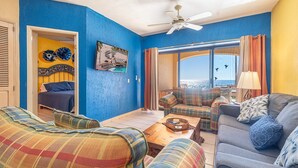 A room with a blue wall, a bed with a headboard, a sofa, a chair, a table, and a view of the sea through a window.